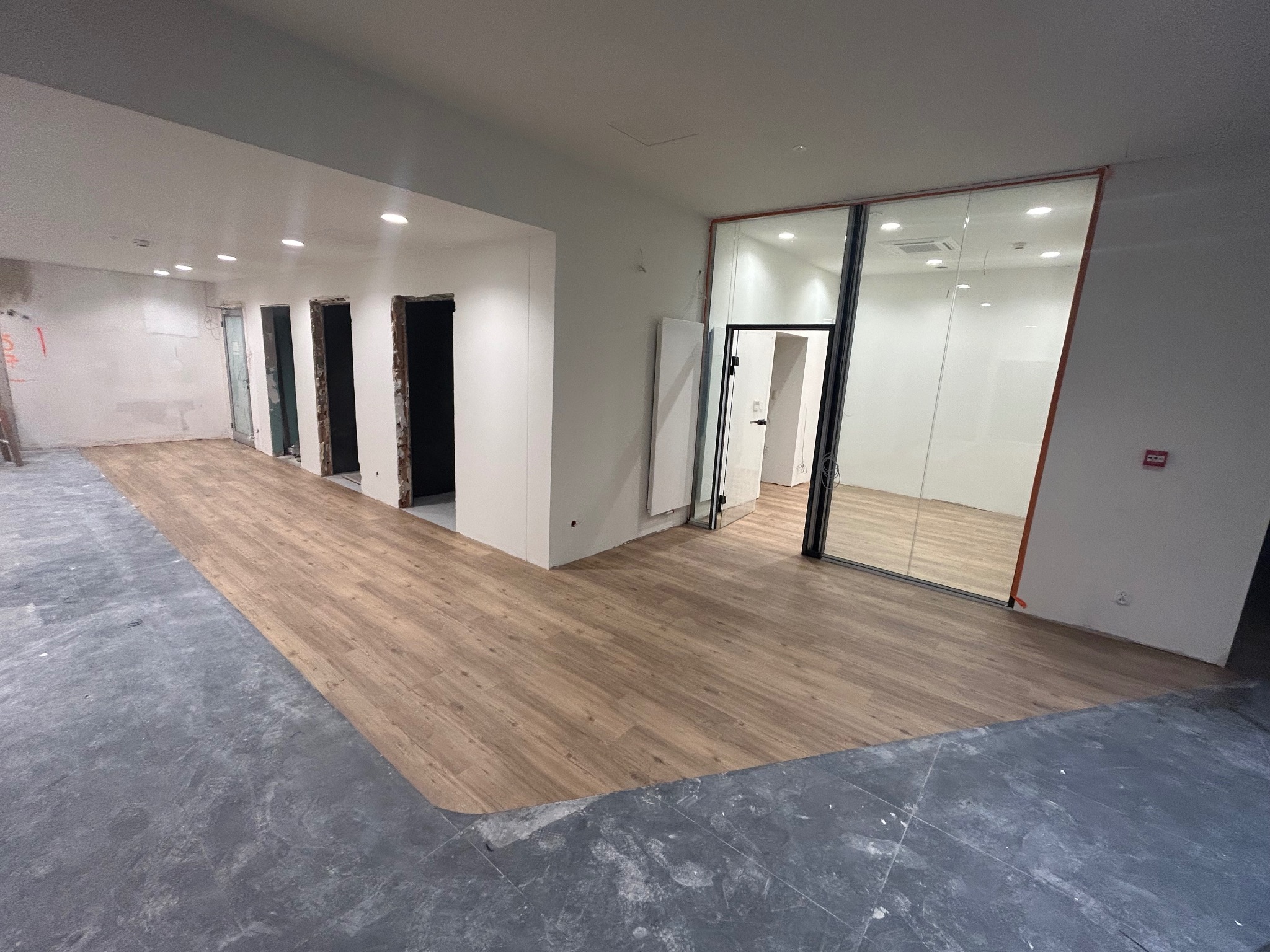 Fragment pomieszczenia w trakcie prac wykończeniowych: ułożone panele podłogowe imitujące drewno kontrastują z surowym, betonowym podłożem. Widoczne otwarte przestrzenie drzwiowe i przeszklone ściany.