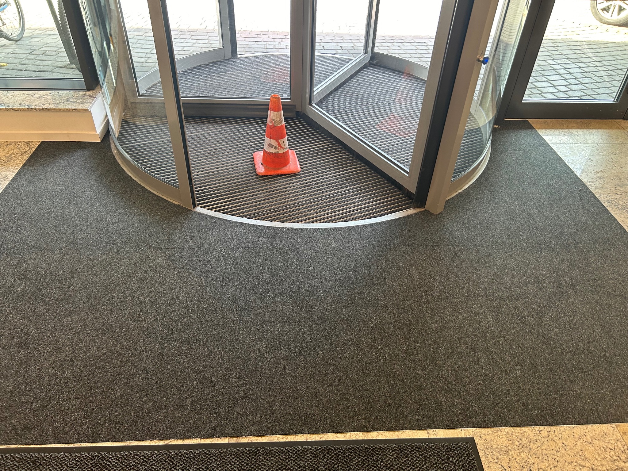 Szara wykładzina dywanowa przy wejściu obrotowym, pomarańczowy stożek ostrzegawczy na metalowej wycieraczce wewnątrz wejścia.