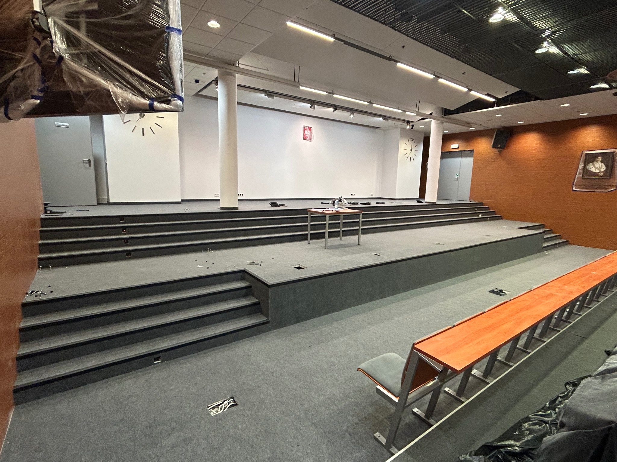 Sala konferencyjna z wielopoziomową podłogą pokrytą szarą wykładziną dywanową, widoczne niedociągnięcia montażowe i elementy instalacji elektrycznej w podłodze, przygotowania do wydarzenia.