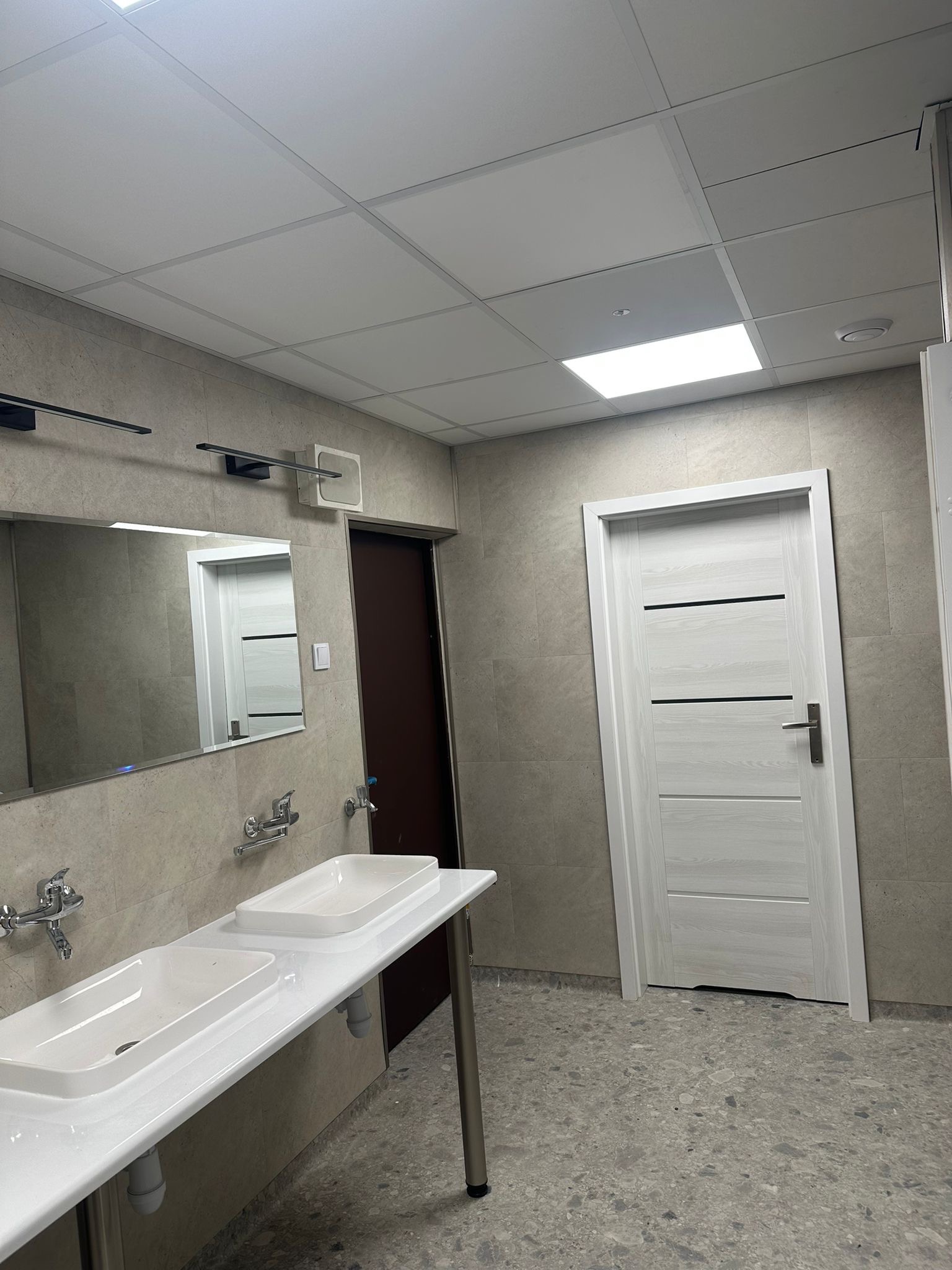 Nowoczesna łazienka publiczna z dwoma białymi umywalkami nablatowymi na metalowych nogach, szarą płytką na ścianach i podłodze, oraz białą, wewnętrzną ościeżnicą drzwiową z poziomymi wstawkami.