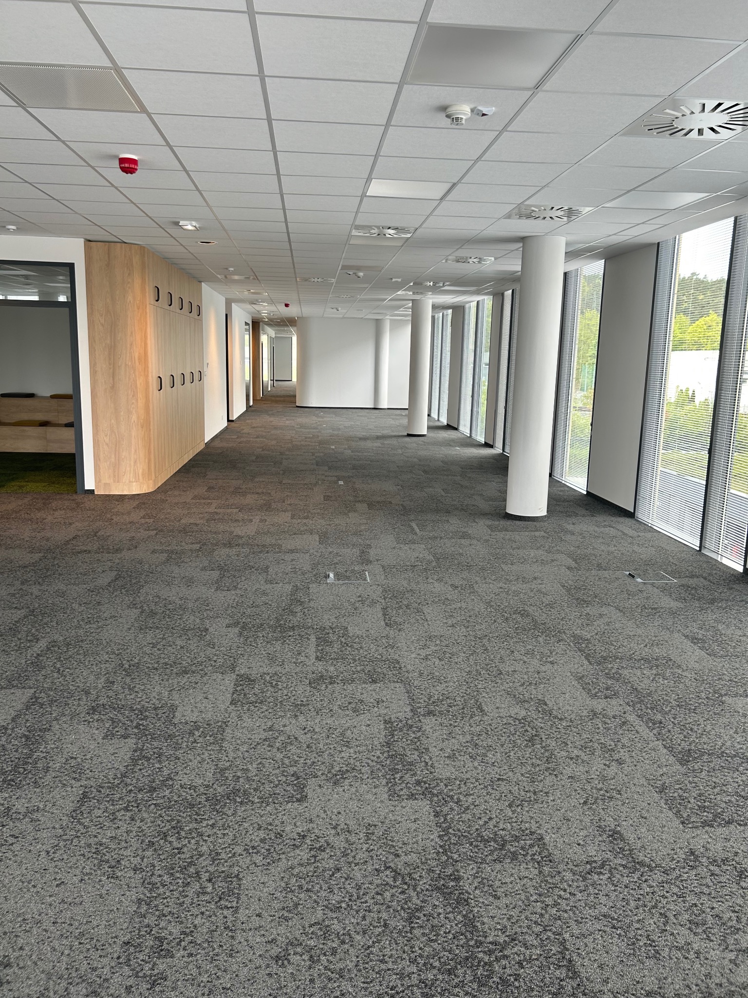 Wnętrze biurowe z szarą, wzorzystą wykładziną dywanową. Długi korytarz z kolumnami i oknami. Widoczne szafki wzdłuż ściany. Wnętrze jasne, oświetlone światłem dziennym.