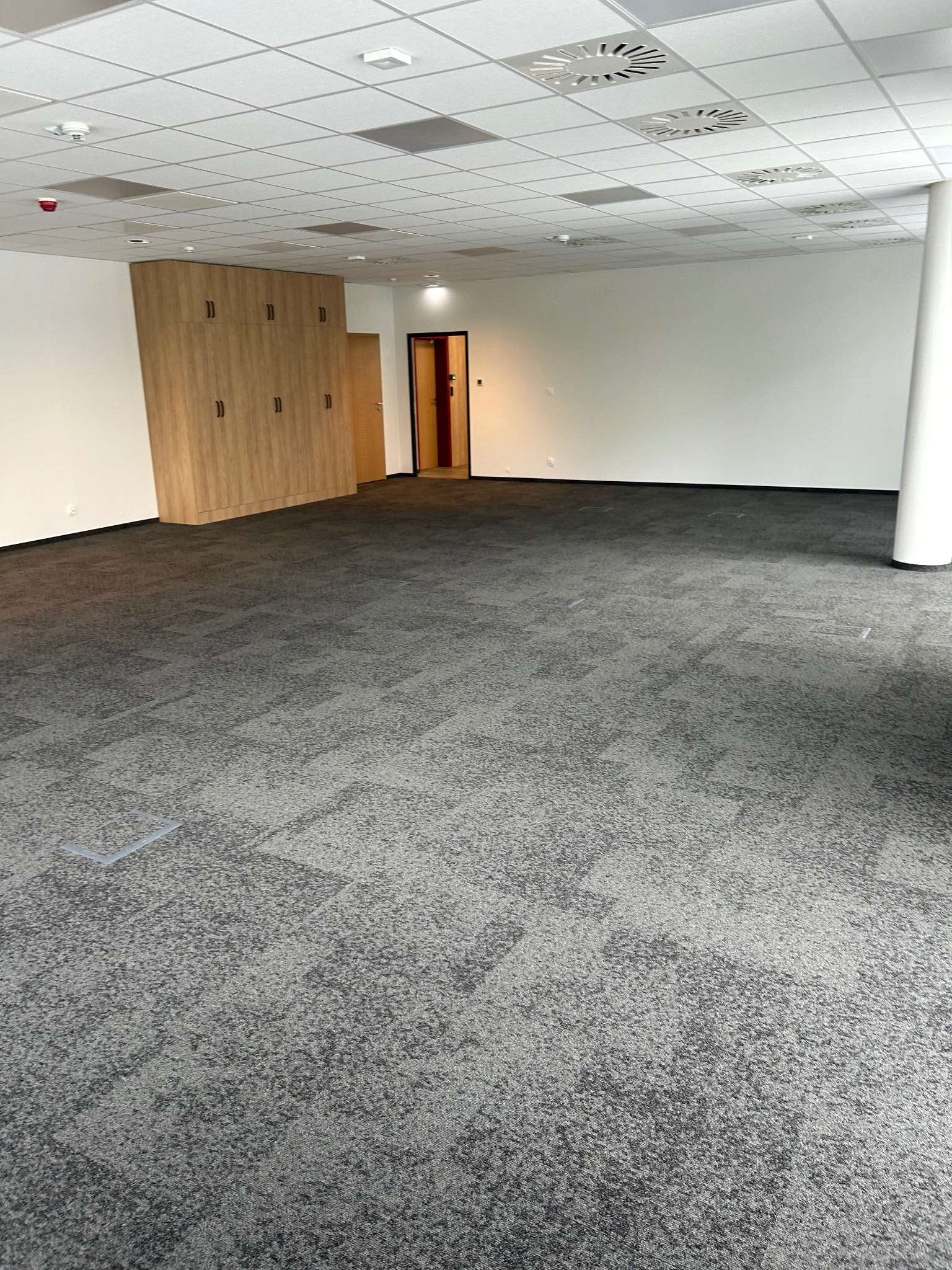Szara wykładzina dywanowa w biurze z drewnianą zabudową i białym sufitem kasetonowym. Widoczne elementy wentylacji i oświetlenia.