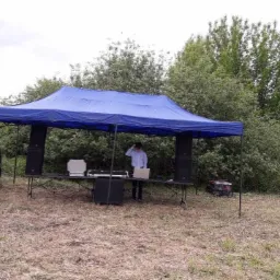 Rozstawiony niebieski namiot z nagłośnieniem i sprzętem DJ-skim na łące, przygotowania do imprezy plenerowej.