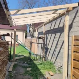 Wznoszenie drewnianej konstrukcji dachu na tle betonowego budynku, widoczne rusztowania i składowane deski.