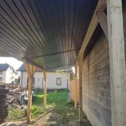 Zadaszenie tarasu z blachy trapezowej w kolorze grafitowym, wsparte na drewnianych słupach, widok z perspektywy ogrodu, w tle budynek mieszkalny.