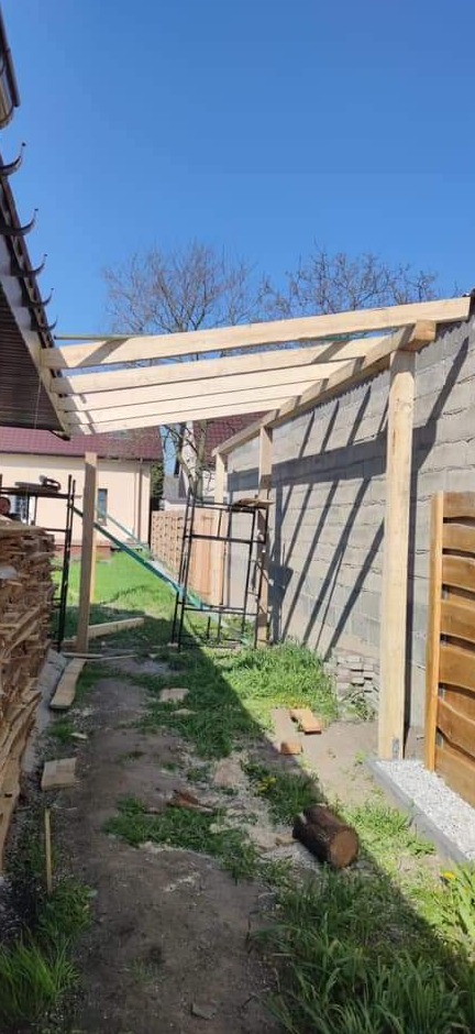 Wznoszenie drewnianej konstrukcji dachu na tle betonowego budynku, widoczne rusztowania i składowane deski.