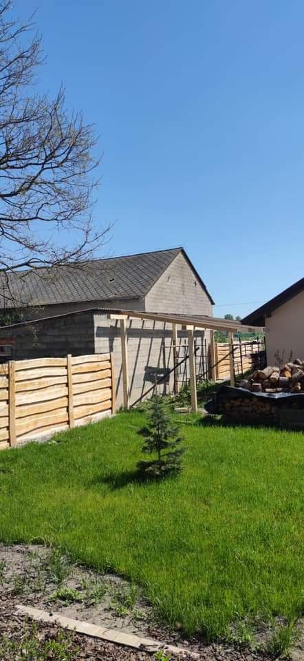 Widok na ogród z niskim drewnianym płotem, w tle budynek z szarym dachem i konstrukcja z drewnianych belek, prawdopodobnie zadaszenie w trakcie budowy.