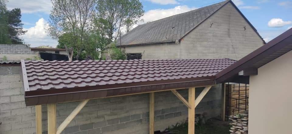 Nowa, brązowa blachodachówka na drewnianej konstrukcji w ogrodzie, na tle starego, murowanego budynku gospodarczego i błękitnego nieba.