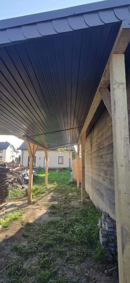Zadaszenie tarasu z blachy trapezowej w kolorze grafitowym, wsparte na drewnianych słupach, widok z perspektywy ogrodu, w tle budynek mieszkalny.