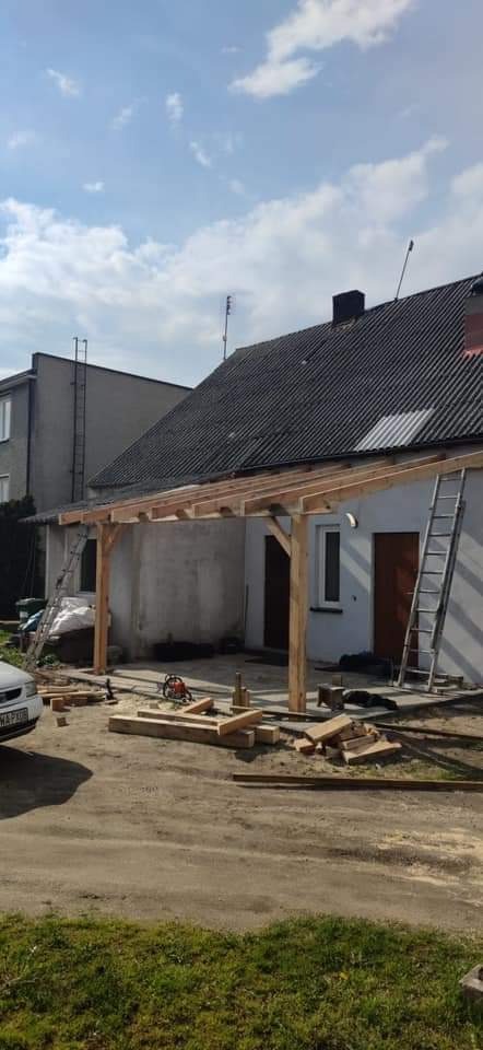 Drewniana konstrukcja zadaszenia tarasu w trakcie budowy, widoczny piła łańcuchowa, resztki drewna i drabina oparta o ścianę budynku.