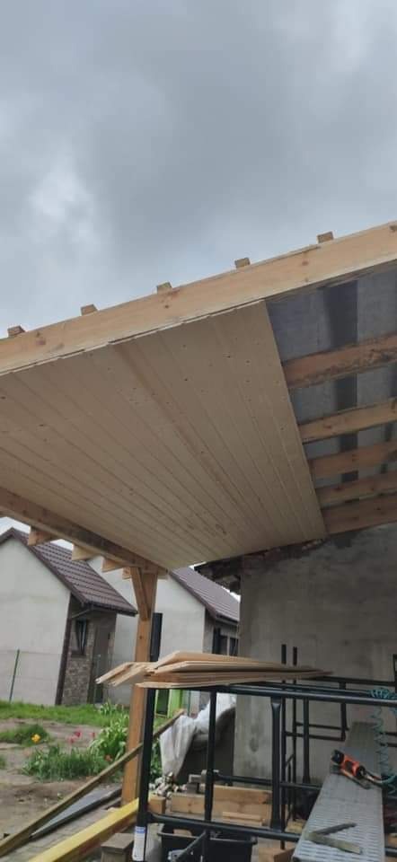 Montaż podbitki dachowej z drewnianych paneli na nowo budowanym zadaszeniu, widoczne elementy więźby dachowej i fragmenty budynków mieszkalnych w tle.