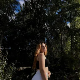 Portret młodej kobiety w czarnym topie i białych spodniach, pozującej na tle leśnej scenerii z wysokimi drzewami i błękitnym niebem w tle, uchwycony w naturalnym świetle.