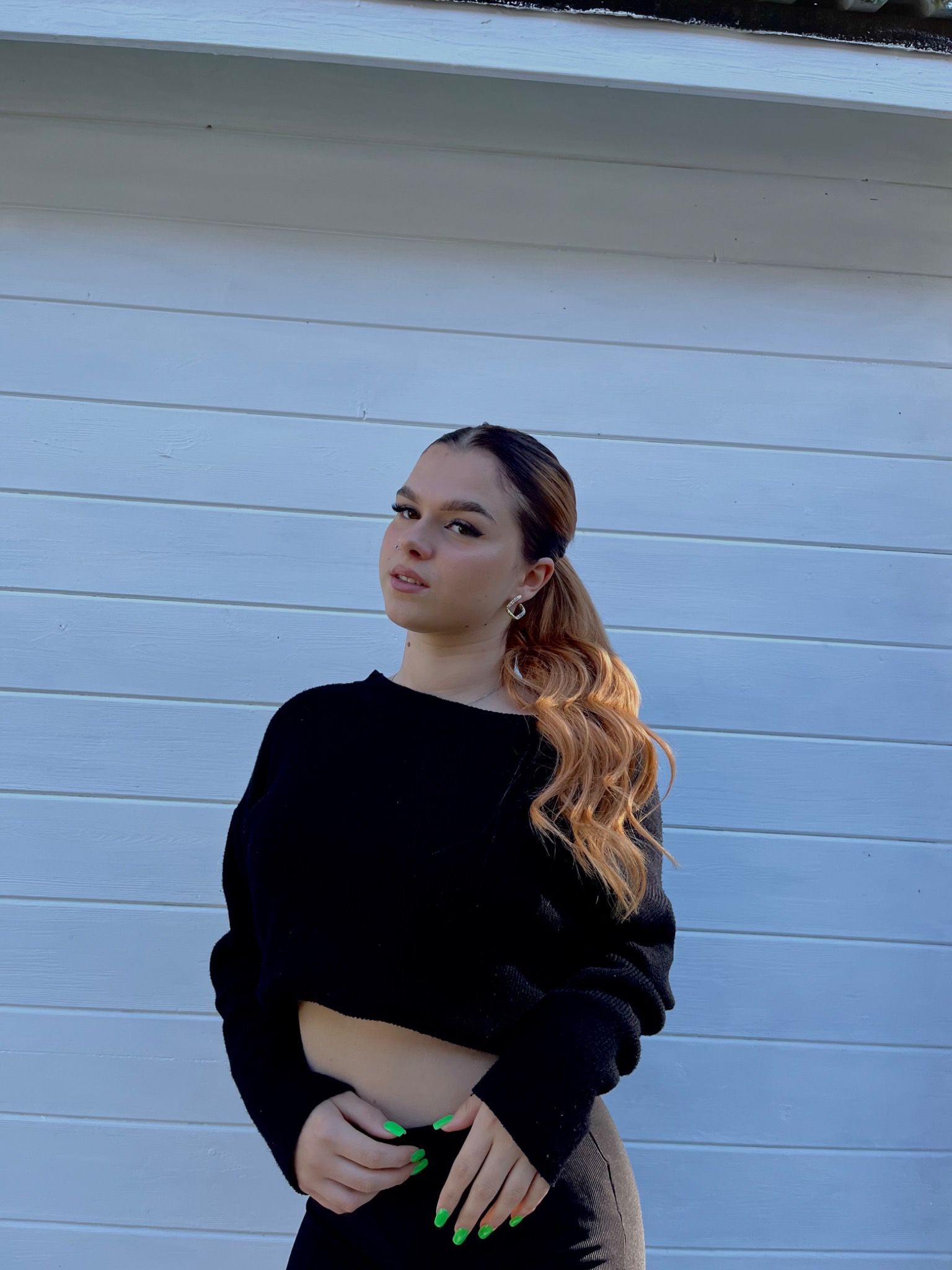 Portret młodej kobiety w czarnym stroju, z włosami upiętymi w kucyk z ombre, na tle białej, drewnianej ściany. Widoczne neonowe paznokcie i srebrne kolczyki.