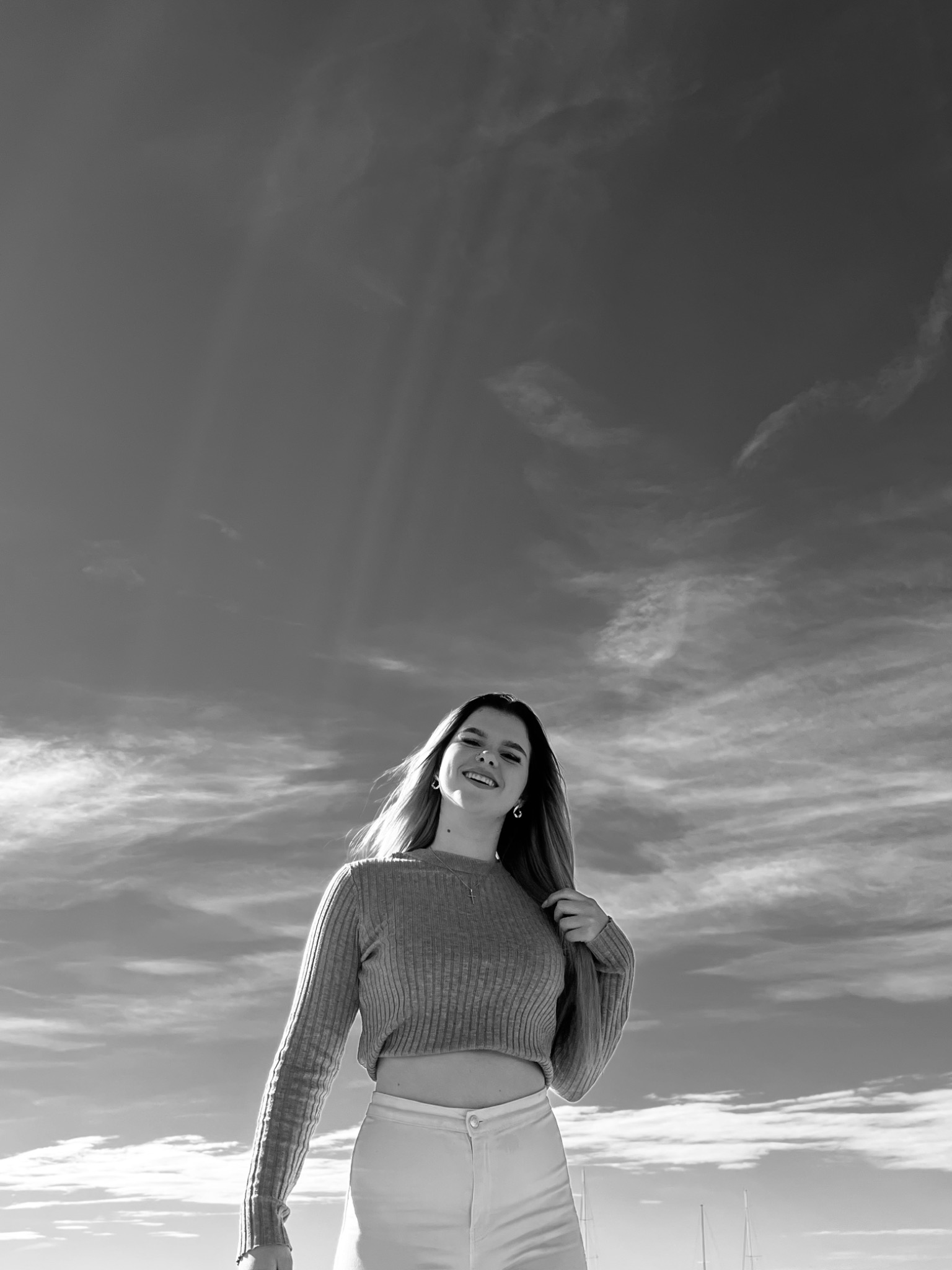 Czarno-białe zdjęcie uśmiechniętej kobiety w swetrze i spodniach na tle pochmurnego nieba z masztami jachtów w oddali.