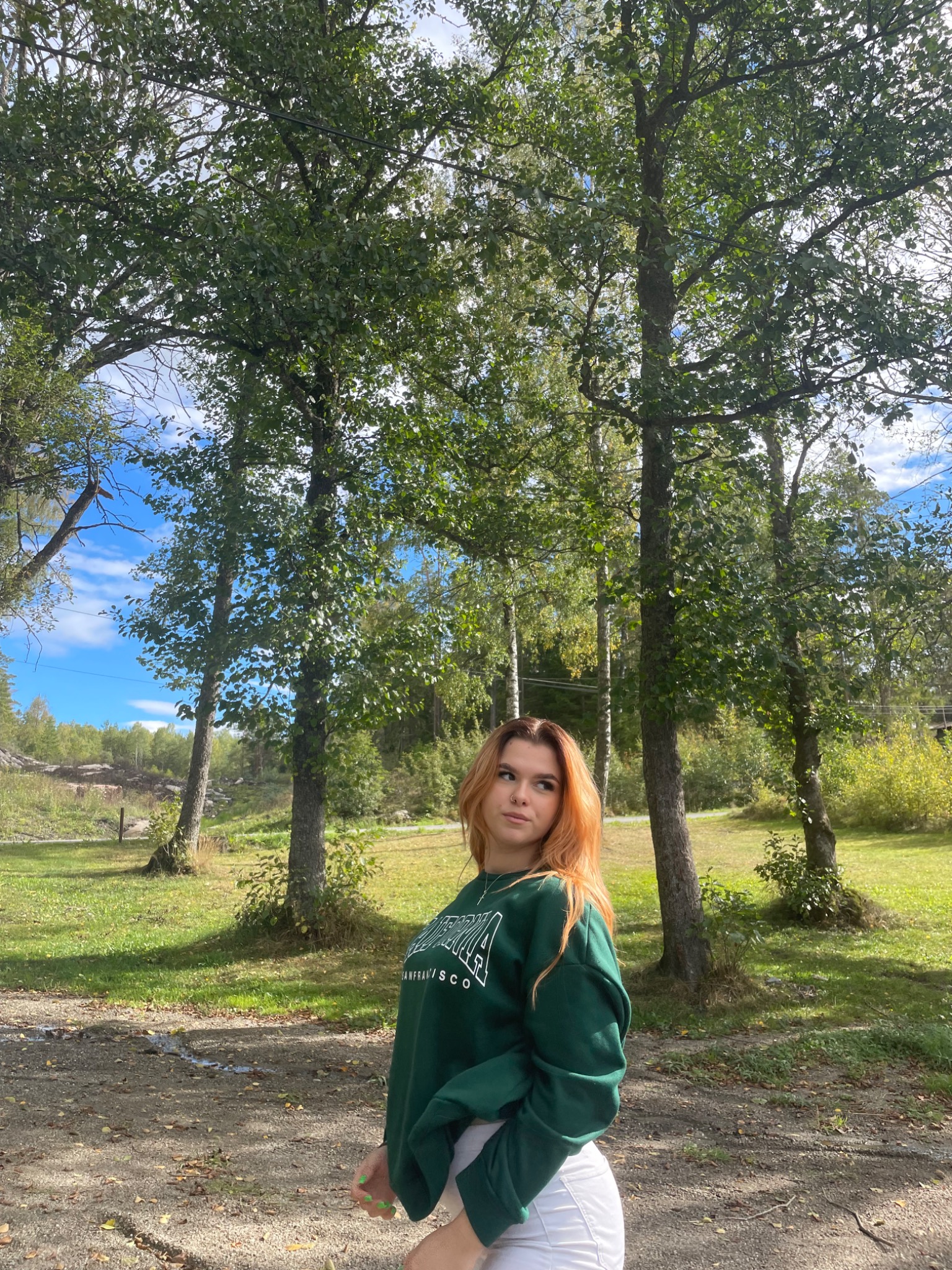 Młoda kobieta z rudymi włosami w zielonej bluzie z napisem San Francisco, pozująca na tle drzew w parku w słoneczny dzień.