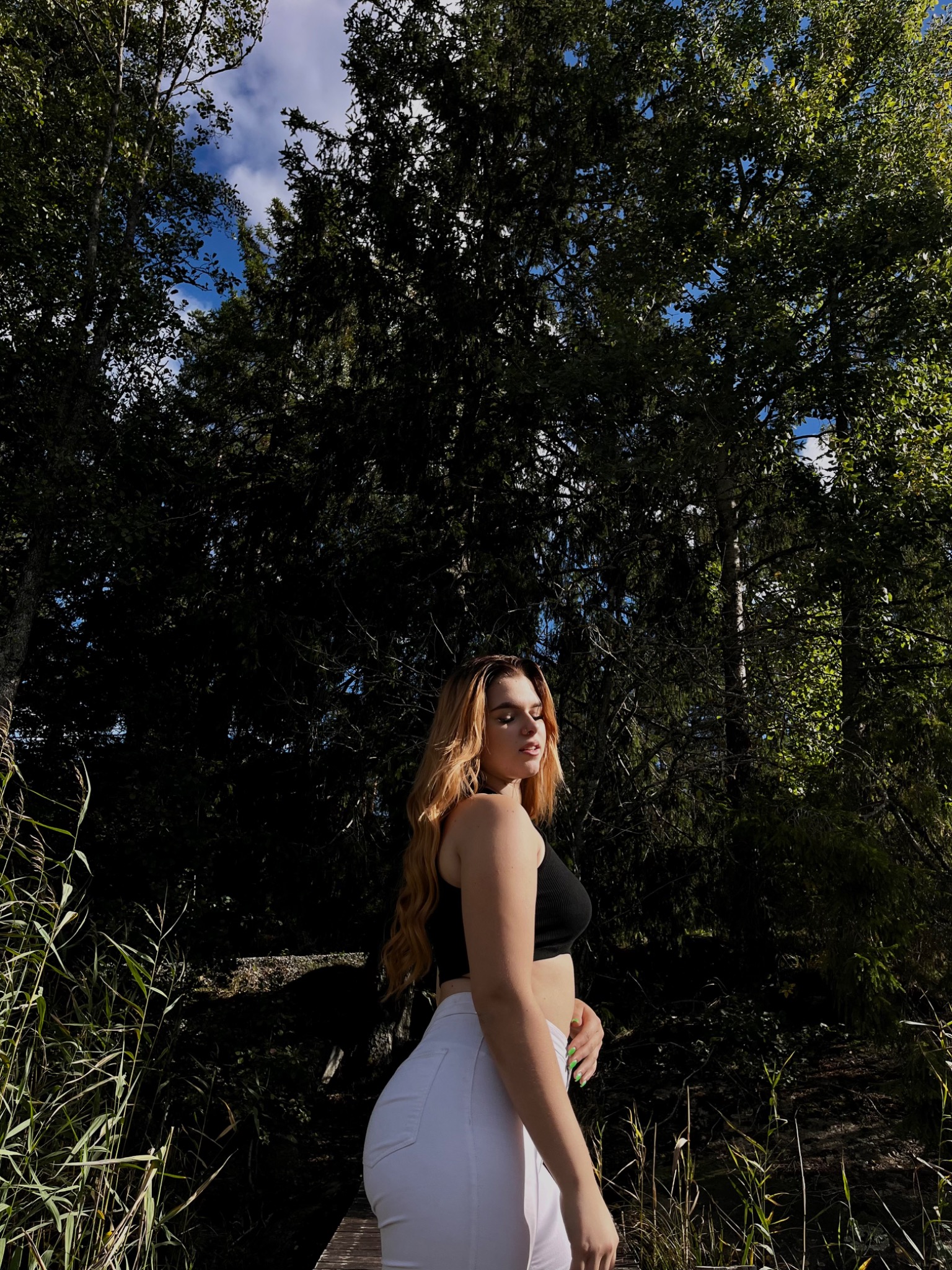 Portret młodej kobiety w czarnym topie i białych spodniach, pozującej na tle leśnej scenerii z wysokimi drzewami i błękitnym niebem w tle, uchwycony w naturalnym świetle.