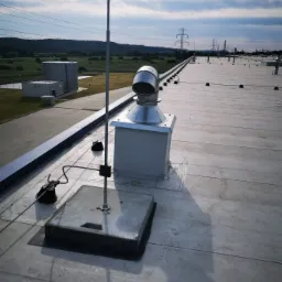 Metalowa instalacja odgromowa z przewodem na płaskim dachu budynku przemysłowego, w tle słupy wysokiego napięcia i zieleń.