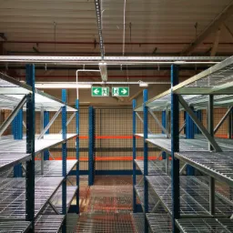 Puste, metalowe regały magazynowe z niebieskimi słupkami, widok z perspektywy korytarza, z zielonymi znakami ewakuacyjnymi pod sufitem.