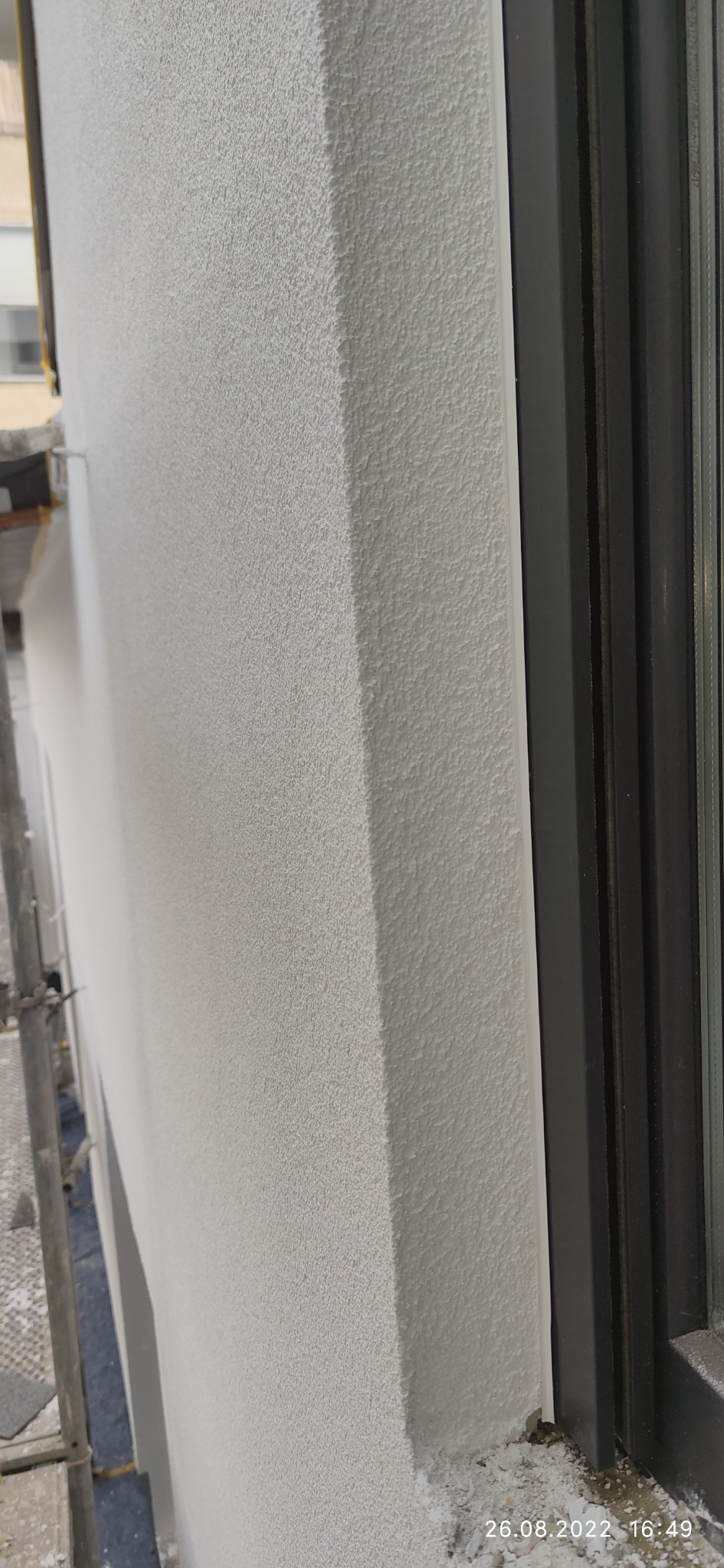 Szara, lekko chropowata elewacja budynku przy ciemnej ramie okiennej, widoczne ślady zaprawy na parapecie.