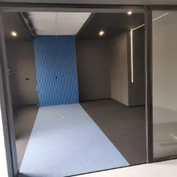 Wnętrze biurowe z niebieską ścianą z paneli, ciemną podłogą i sufitem oraz ledowym oświetleniem liniowym, widok przez przesuwne szklane drzwi.