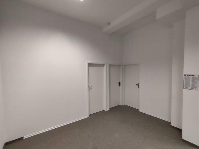 Pomalowane na biało wnętrze biura z trzema białymi drzwiami i szarą wykładziną podłogową; na ścianie po prawej stronie wisi tabliczka informacyjna.