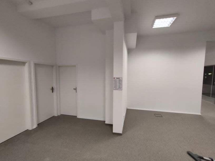 Wnętrze biurowe po odświeżeniu, widoczne białe ściany, szara wykładzina, trzy białe drzwi i oświetlenie sufitowe