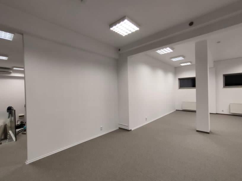 Pomalowane na biało, puste wnętrze biurowe z szarą wykładziną, widok z niskiego kąta, oświetlenie sufitowe, grzejniki pod oknami.