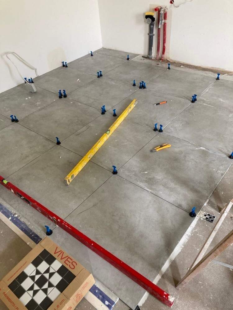 Szare płytki podłogowe w trakcie układania, z niebieskimi klipsami poziomującymi, żółtą poziomicą, czerwoną łatą i kartonem płytek VIVES w rogu pomieszczenia.