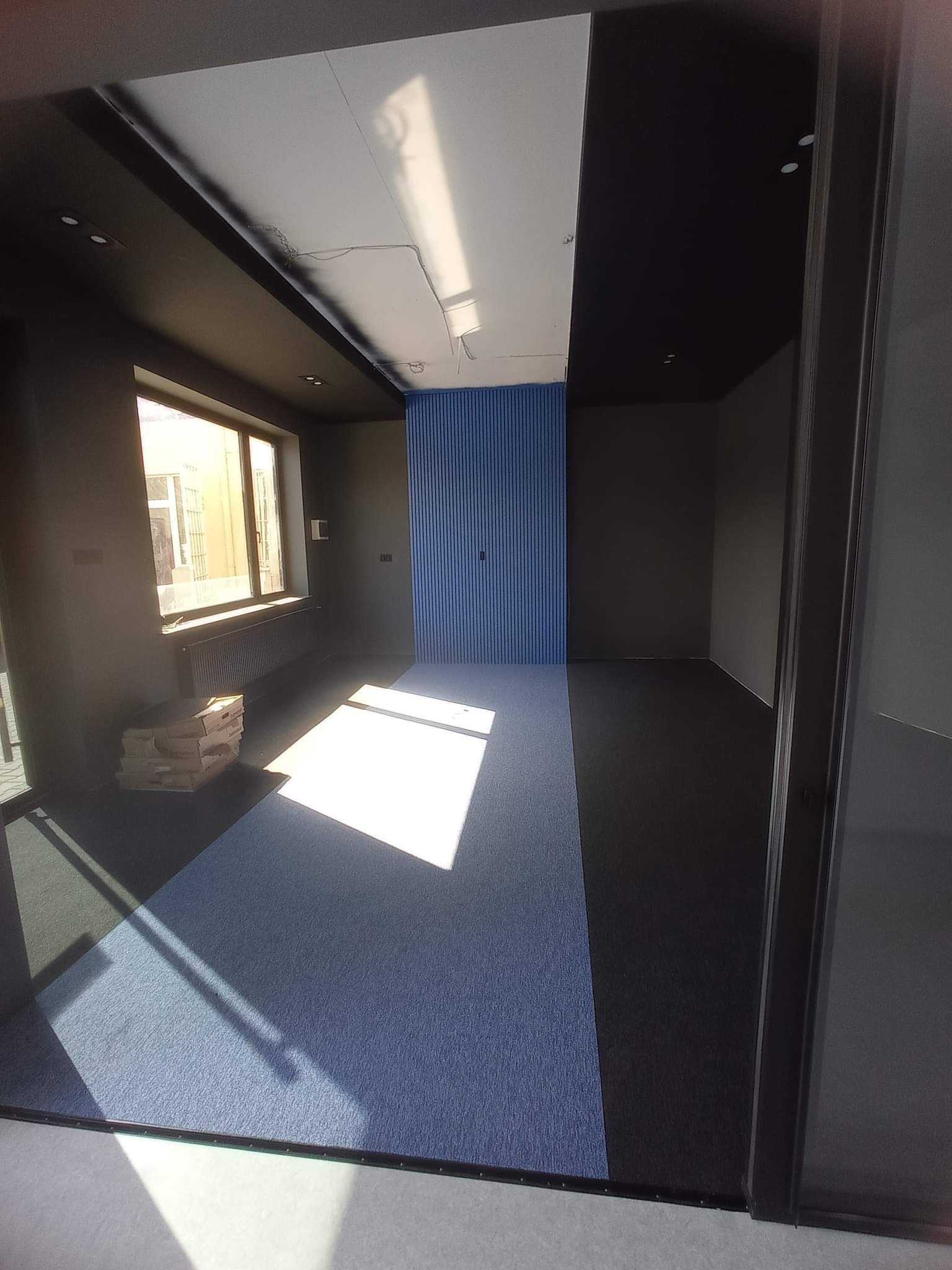 Pusta przestrzeń biurowa z nową podłogą, pomalowanymi ścianami na ciemny kolor i niebieskim akcentem na ścianie, w rogu stos kartonów z panelami podłogowymi, widok na zewnątrz przez okno.