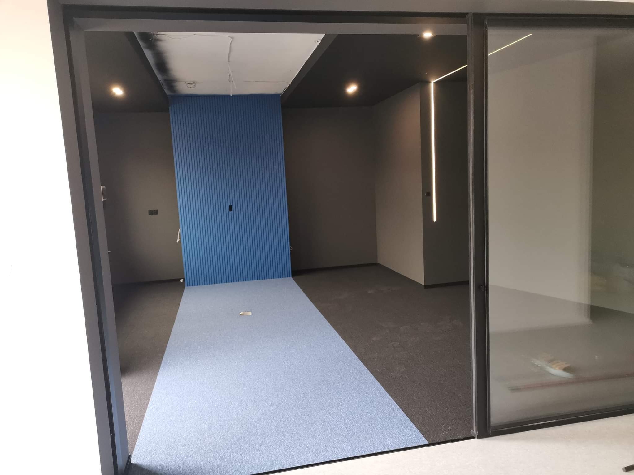 Wnętrze biurowe z niebieską ścianą z paneli, ciemną podłogą i sufitem oraz ledowym oświetleniem liniowym, widok przez przesuwne szklane drzwi.