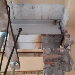 Wsparcie metalowej belki stropowej za pomocą stempli budowlanych, widoczne ślady spawania na belce i fragmenty muru z cegły