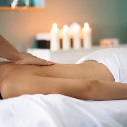 Masaż relaksacyjny pleców w spa, dłoń masażysty na ciele klienta, w tle zapalone świece i kwiaty, miękkie światło, białe prześcieradło.