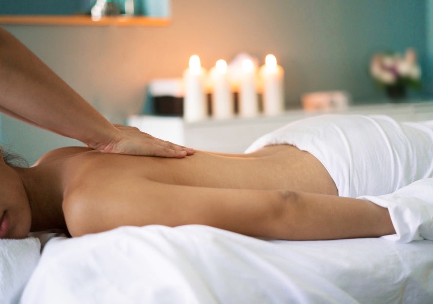 Masaż relaksacyjny pleców w spa, dłoń masażysty na ciele klienta, w tle zapalone świece i kwiaty, miękkie światło, białe prześcieradło.