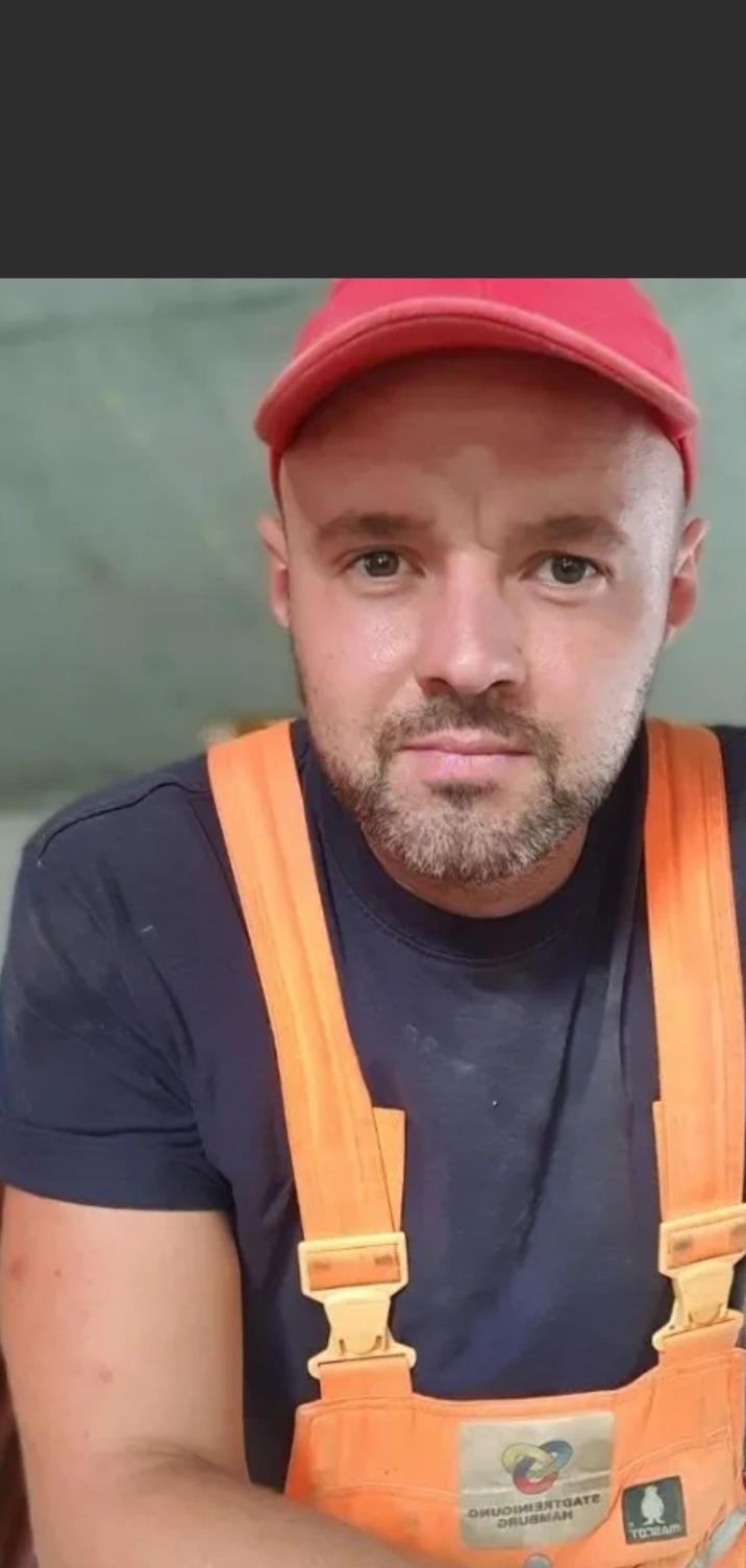 Portret mężczyzny w czerwonej czapce i pomarańczowych szelkach roboczych na tle szarej ściany.