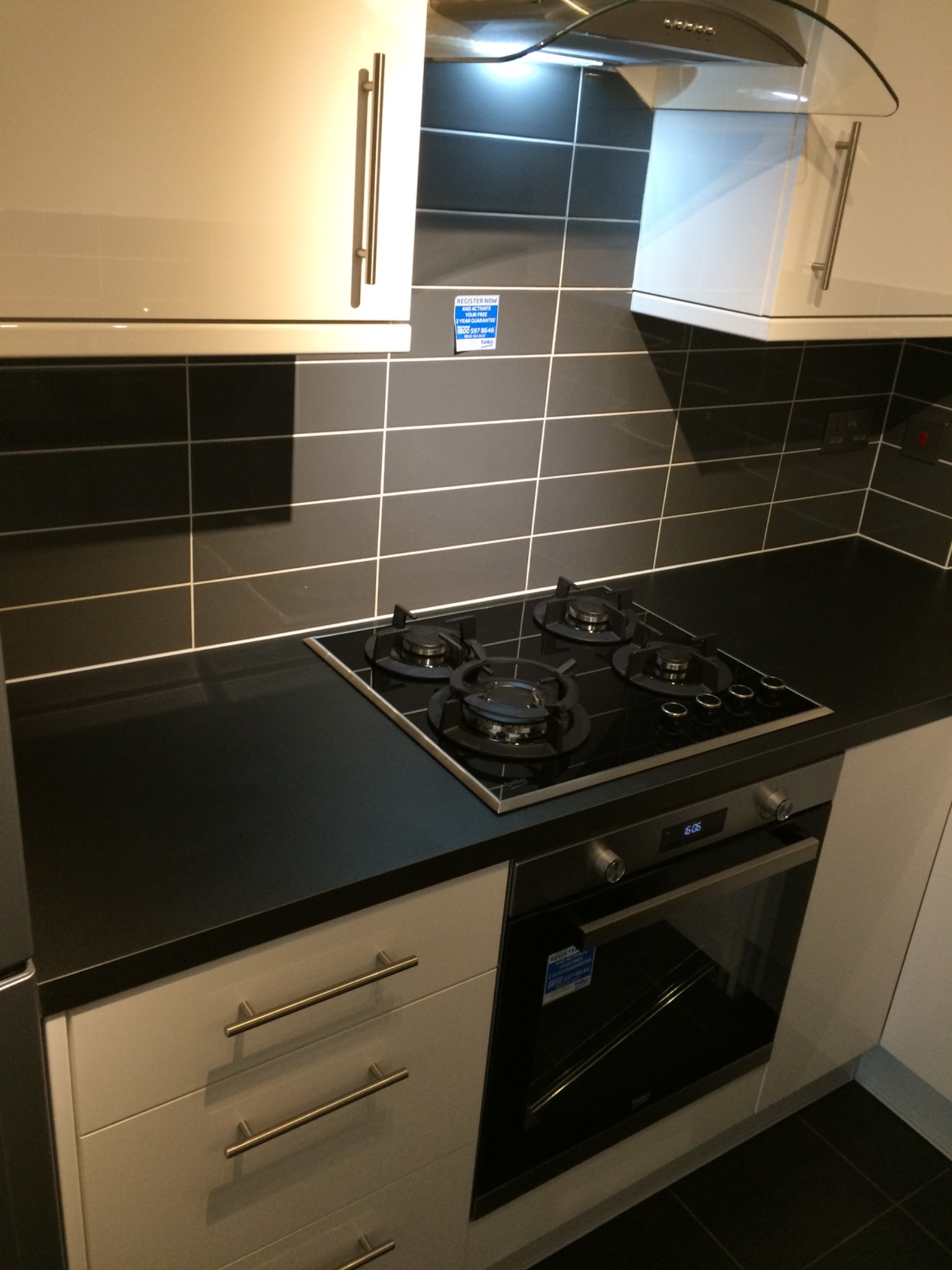 Nowoczesna kuchnia z czarnym blatem, płytą gazową i piekarnikiem, wykończona szarymi płytkami i białymi szafkami z podłużnymi uchwytami.