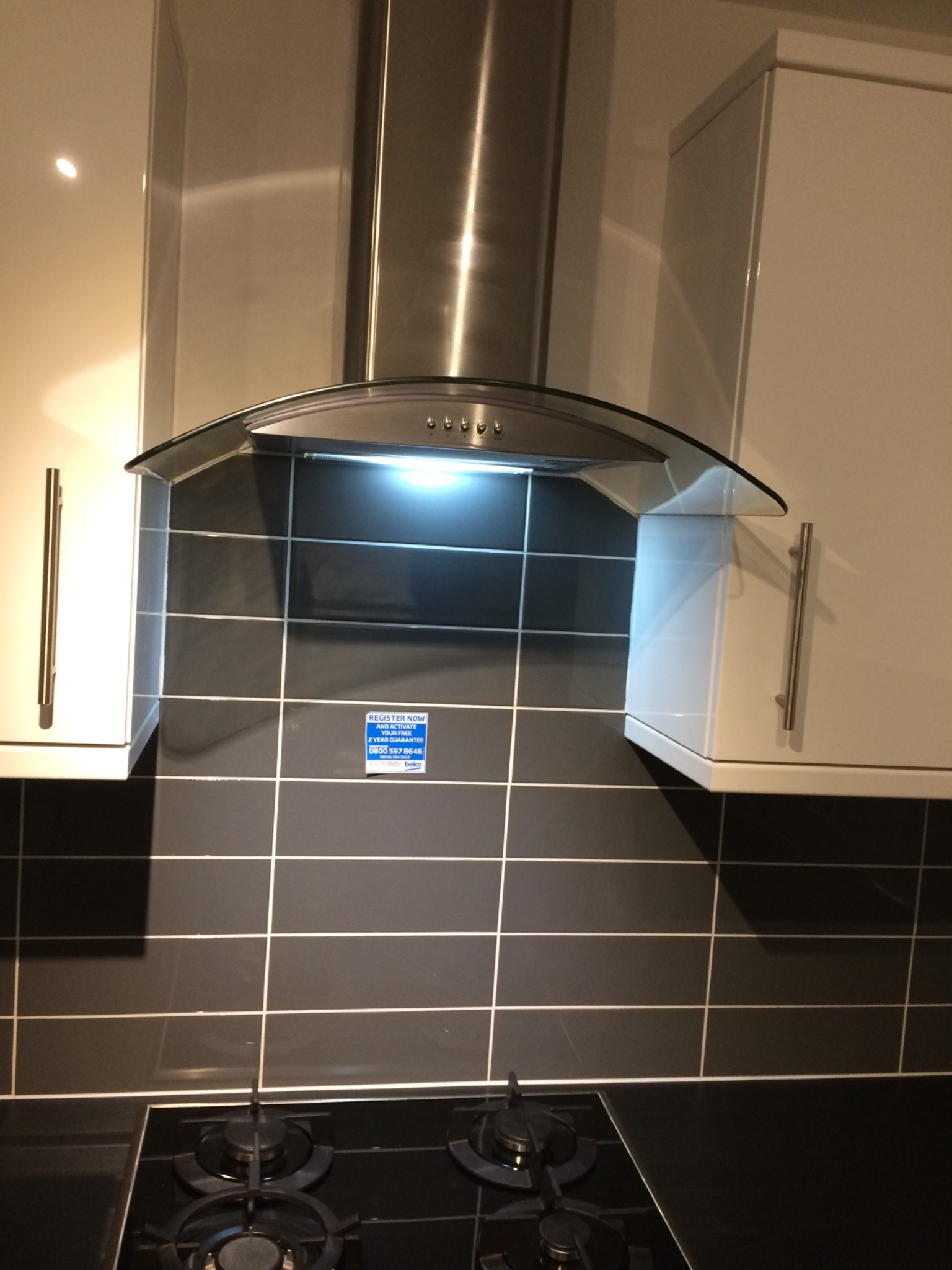 Nowoczesna kuchnia z czarnymi błyszczącymi płytkami ułożonymi w kwadratową siatkę, srebrnym okapem z szklanym daszkiem i białymi szafkami z podłużnymi uchwytami, nad czarną płytą gazową.