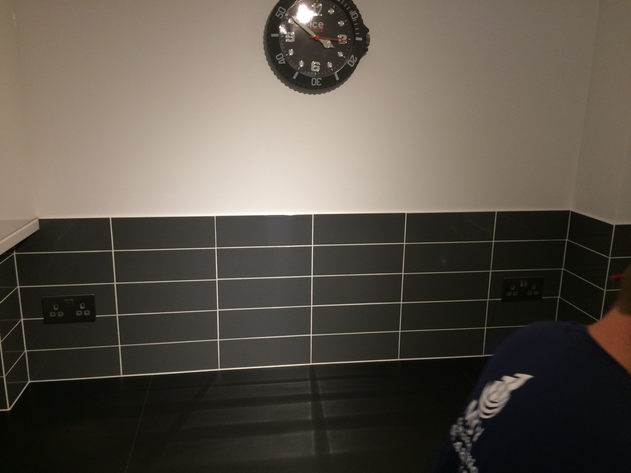 Ściana wyłożona czarnymi płytkami z białymi fugami, nad blatem. Widoczny zegar i gniazdka elektryczne. Fragment sylwetki osoby w prawym dolnym rogu.
