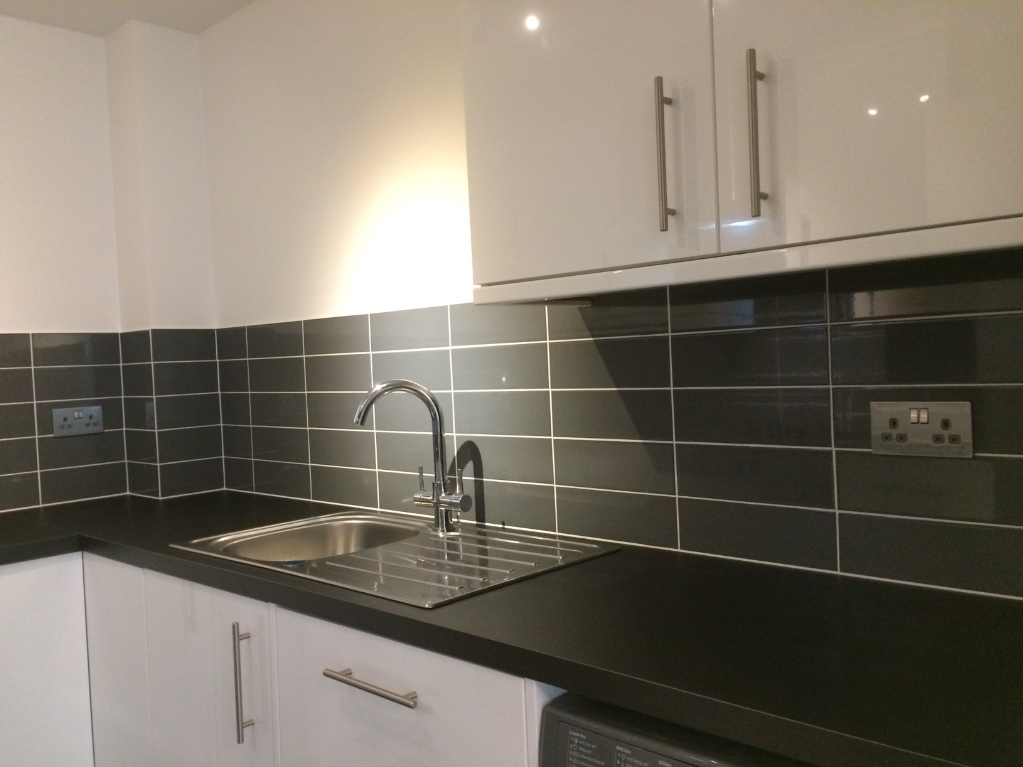 Białe szafki kuchenne z błyszczącymi frontami, czarny blat i czarne płytki ścienne z jasną fugą oraz stalowy zlew z baterią.
