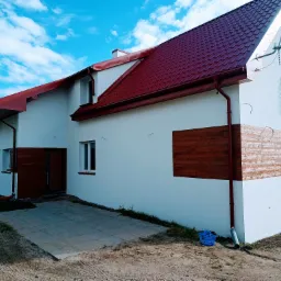 Dwupiętrowy dom z białą elewacją, czerwoną dachówką i drewnianymi panelami dekoracyjnymi na elewacji, widziany z perspektywy ziemi, z niebieskim koszem na pierwszym planie.