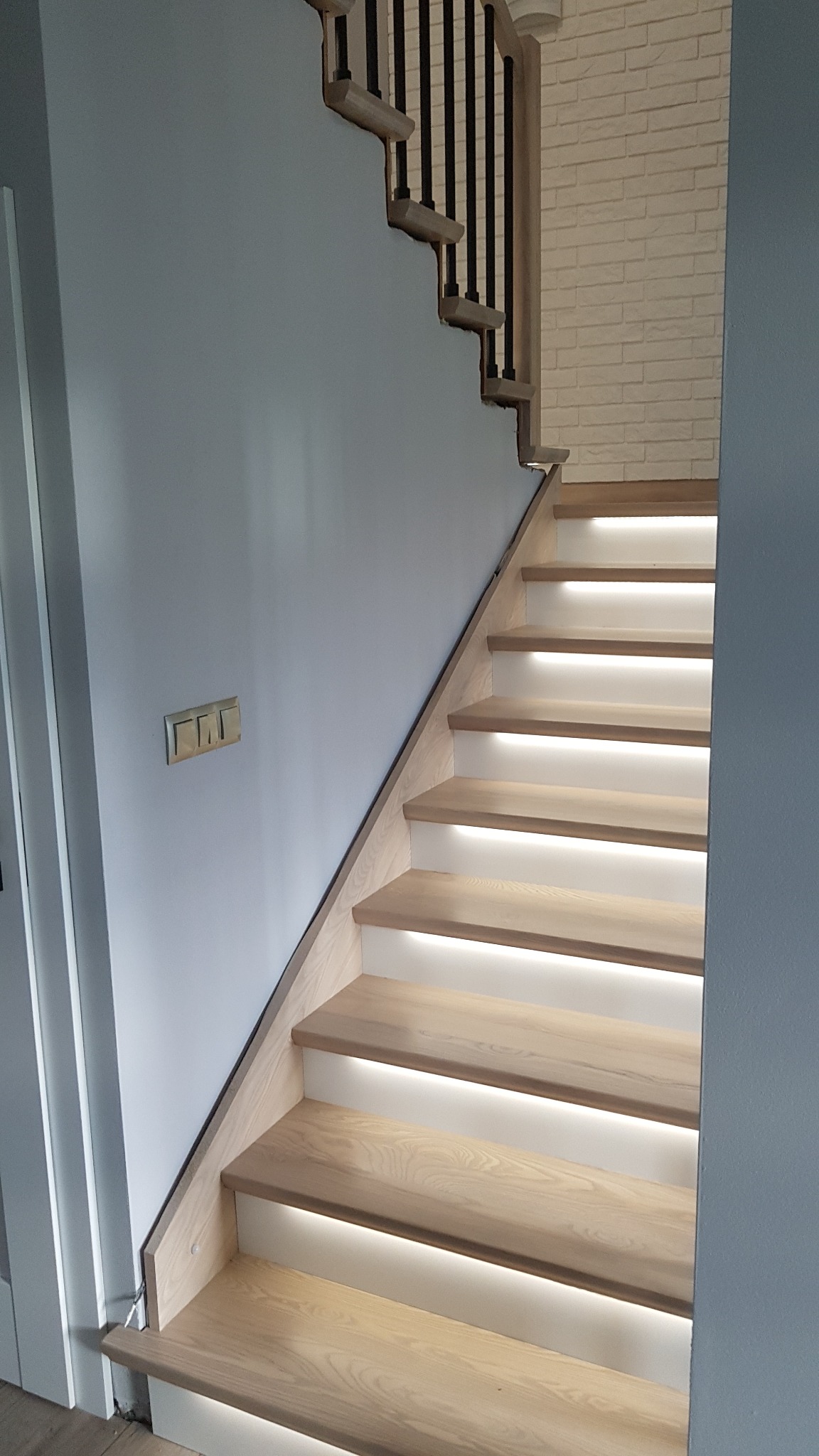 Nowoczesne schody drewniane z podświetleniem LED, czarne metalowe balustrady, ściana pomalowana na szaro, widoczne włączniki elektryczne.