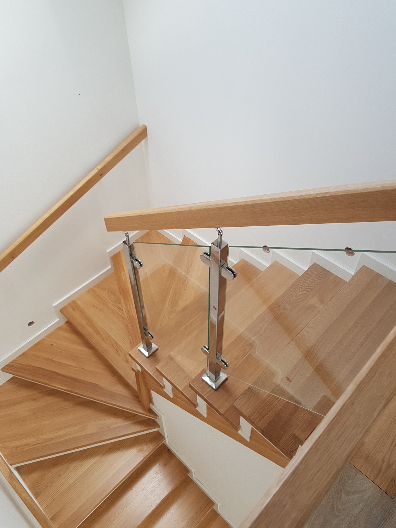 Kątowe ujęcie z góry na drewniane schody z jasnego drewna, z przezroczystą szklaną balustradą mocowaną na chromowanych słupkach, prowadzące w dół w białym wnętrzu.