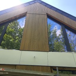 Stanisław kulik - Nowoczesna elewacja budynku z drewnianą okładziną i dużymi oknami z refleksami drzew. Widok z dołu, podkreślający geometrię budynku.