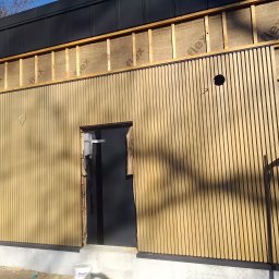 Stanisław kulik - Montaż elewacji z drewnianych listew na budynku. Widoczne elementy konstrukcyjne i izolacja. Częściowo zamontowane listwy tworzą pionowy wzór.