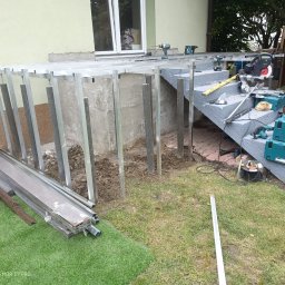 Stanisław kulik - Montaż konstrukcji tarasu: aluminiowe profile, deski kompozytowe obok. Widoczne narzędzia na schodach. Etap budowy tarasu przy domu z zielonym trawnikiem.