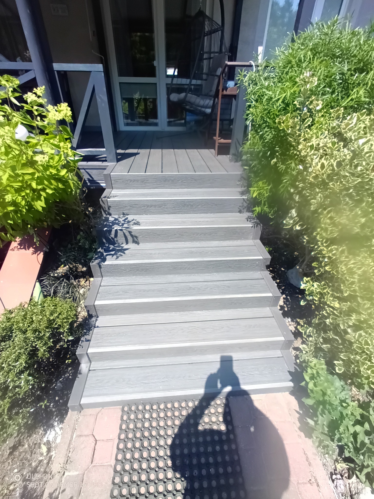 Schody z deski kompozytowej prowadzące do domu, otoczone zielenią. Widoczny fragment tarasu i wejścia do budynku.