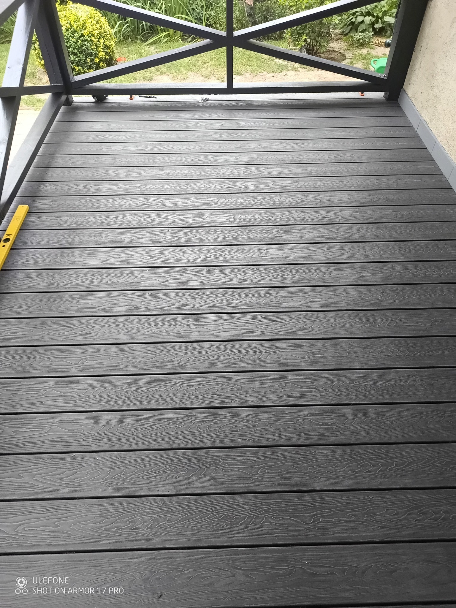 Szary taras kompozytowy z widocznym poziomem na lewej krawędzi, ukazujący precyzję montażu. W tle zieleń ogrodu.