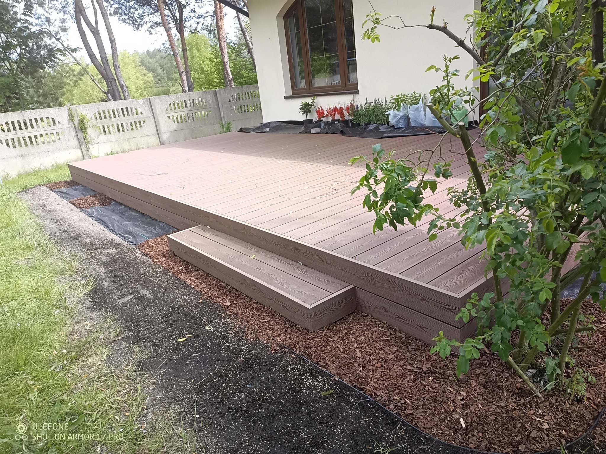 Nowoczesny taras z deski kompozytowej w odcieniach brązu, z niskimi schodkami i obrzeżem z kory, otoczony zielenią. Widok na dom z oknem i betonowe ogrodzenie.