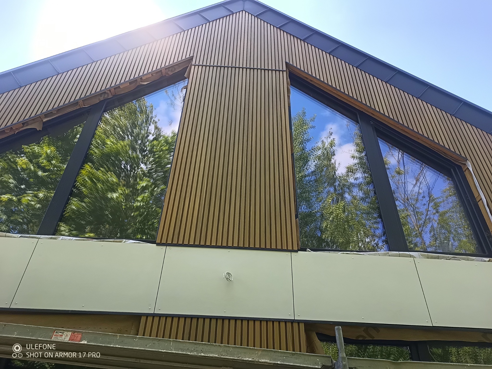 Nowoczesna elewacja budynku z drewnianą okładziną i dużymi oknami z refleksami drzew. Widok z dołu, podkreślający geometrię budynku.