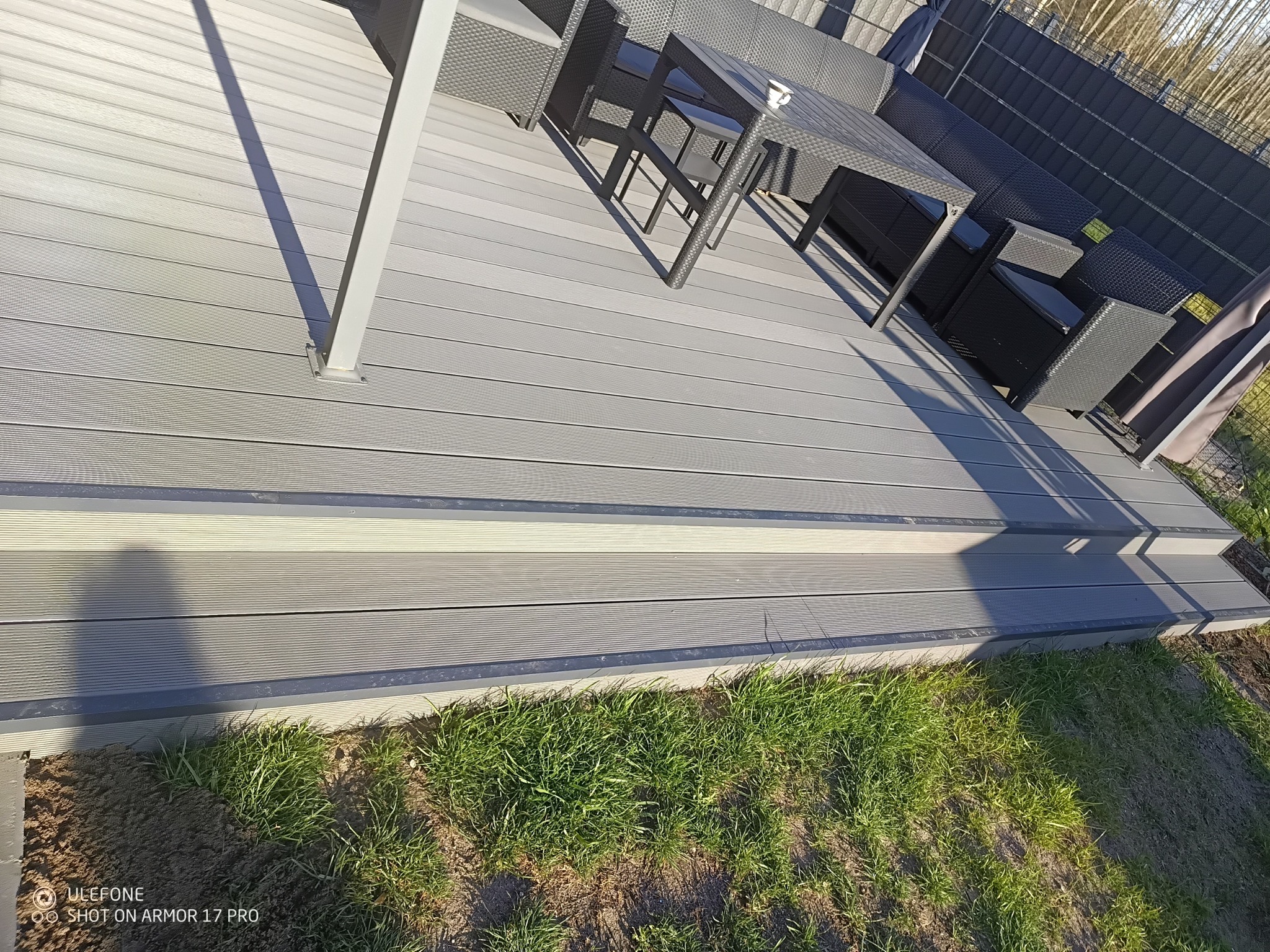 Nowoczesny taras kompozytowy z szarymi deskami, meblami ogrodowymi i schodami, otoczony zielenią. Ujęcie z bliska pokazuje strukturę desek i detale wykończenia.