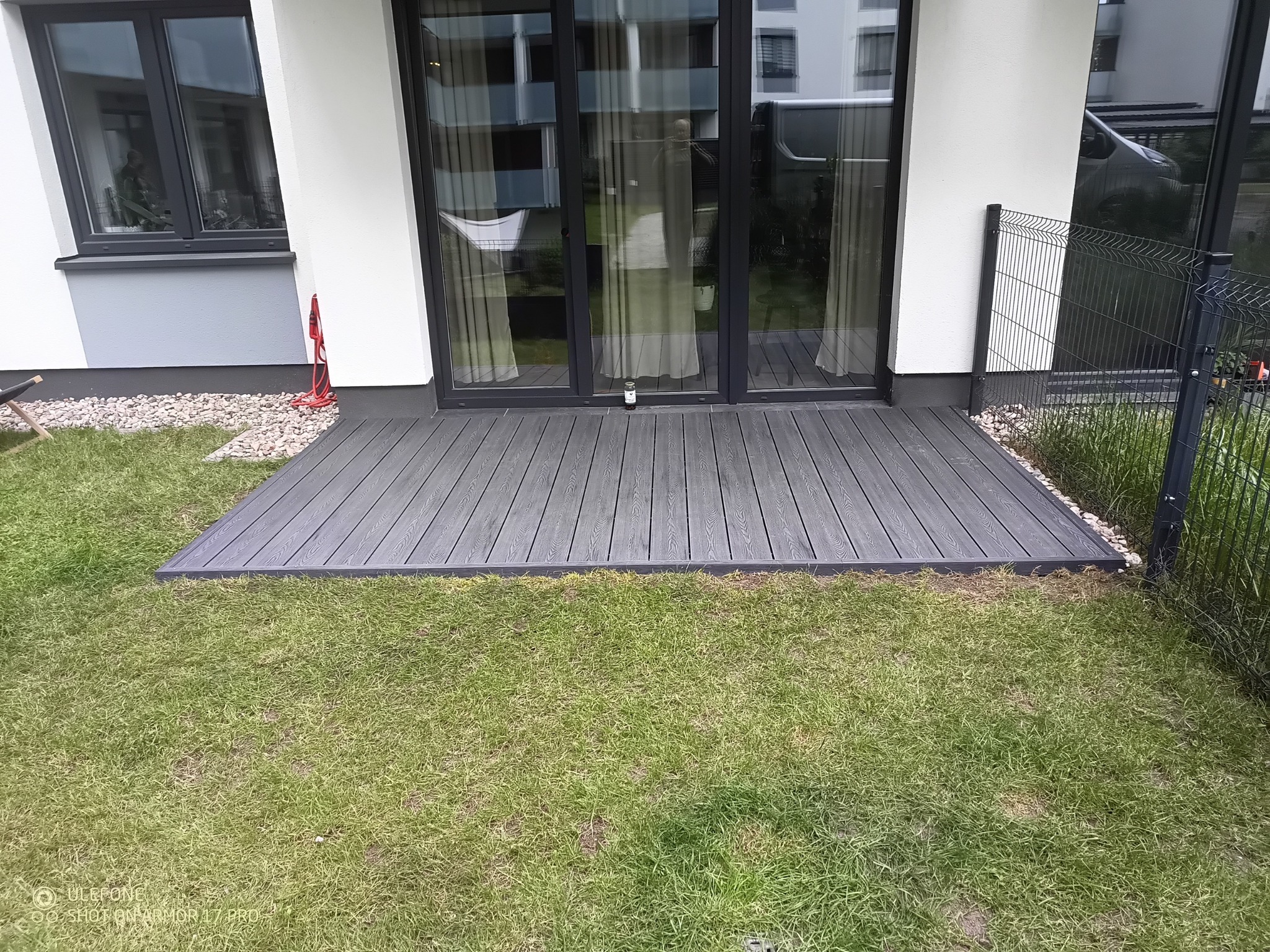 Nowoczesny, szary taras kompozytowy przylegający do domu, z widoczną trawą i elementami ogrodzenia. Minimalistyczny design, idealny na relaks na świeżym powietrzu.